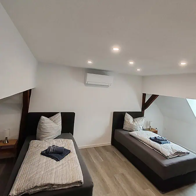 Schlafzimmer mit zwei Einzelbetten, hellen Wänden und modernen Deckenleuchten, ausgestattet mit Kissen und Decken.