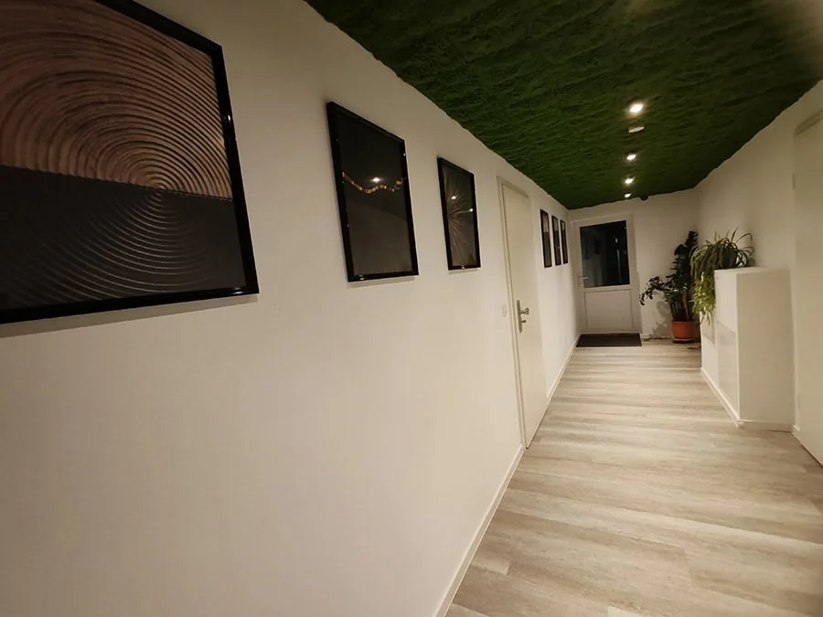 Beleuchteter Flur mit mehreren gerahmten Bildern an der Wand und einem Pflanzengefäß am Ende des Raumes.
